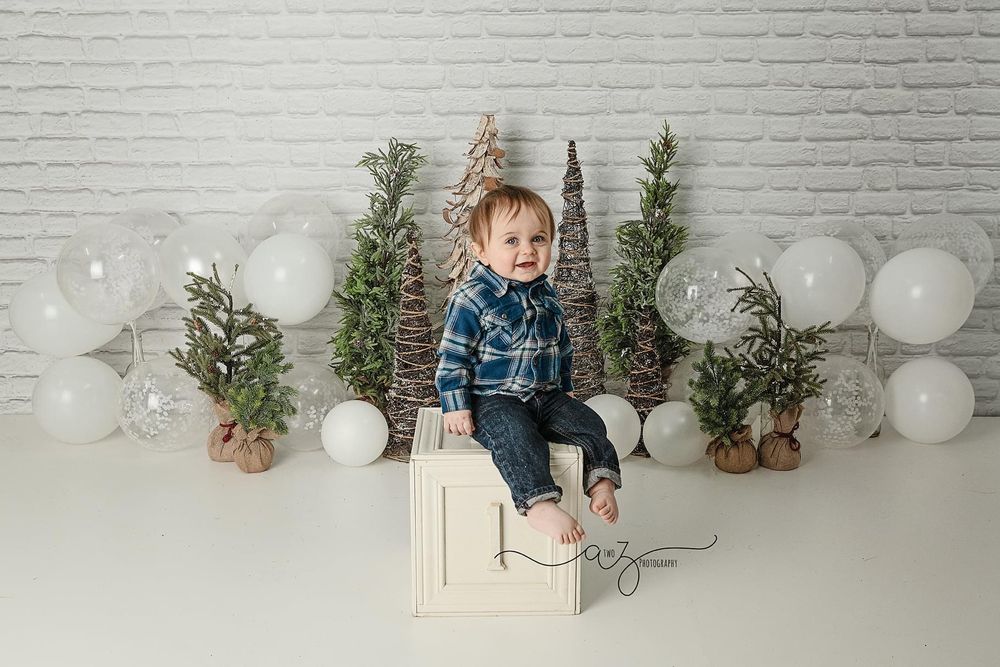 Winter photoshoot setup with greenery, decorative trees, white balloons and a white bench against brick wall.