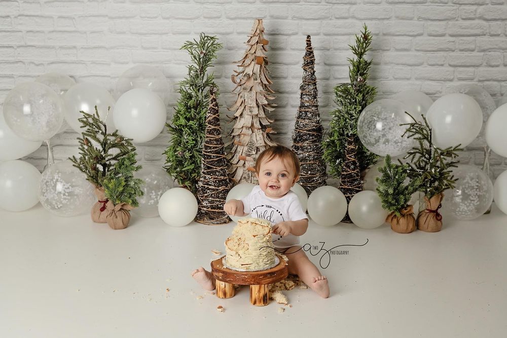 Winter themed photo setup with cake, rustic trees and white balloons against brick wall background.