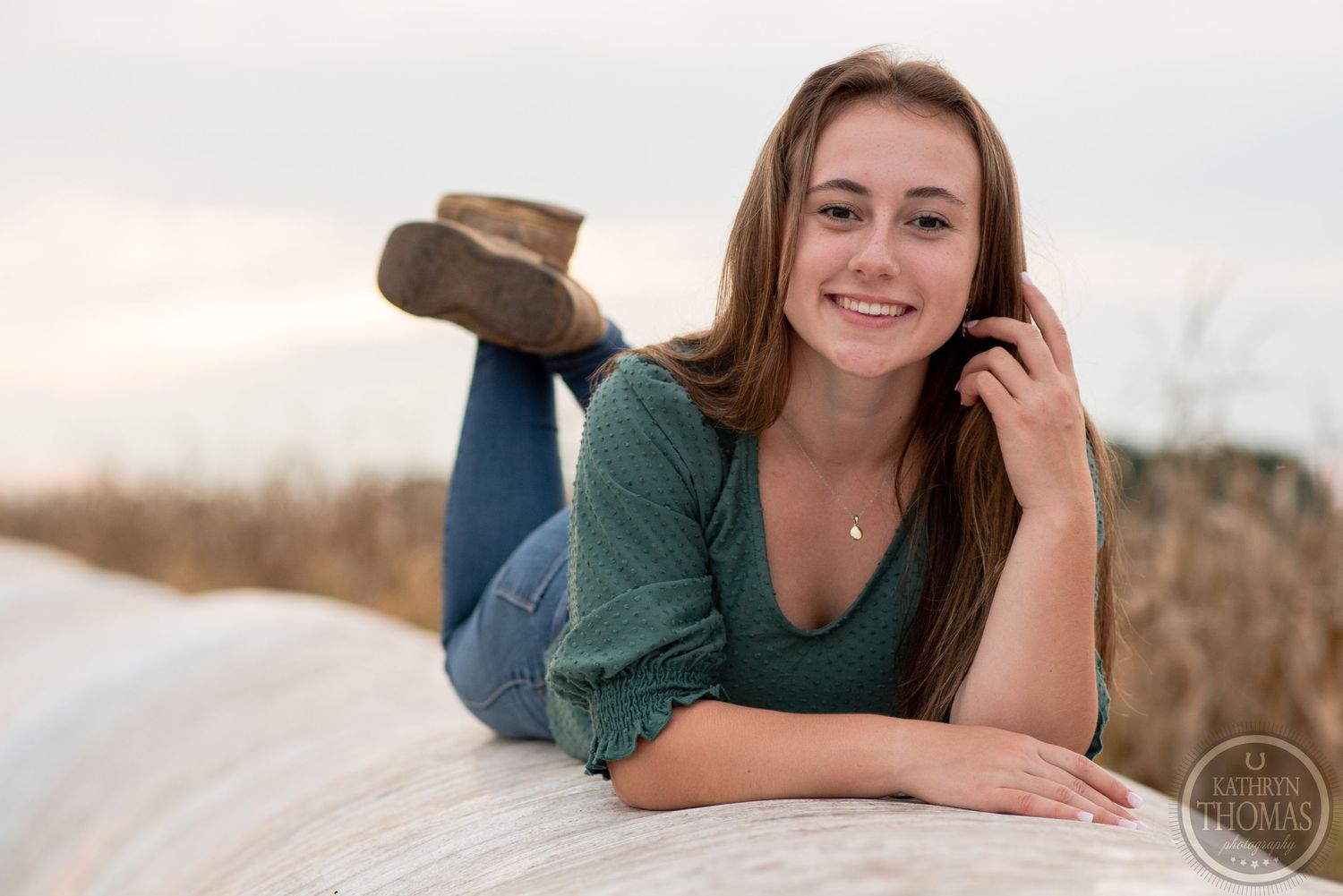 Casual portrait of someone reclining on a hay bale wearing a green top and jeans at sunset.