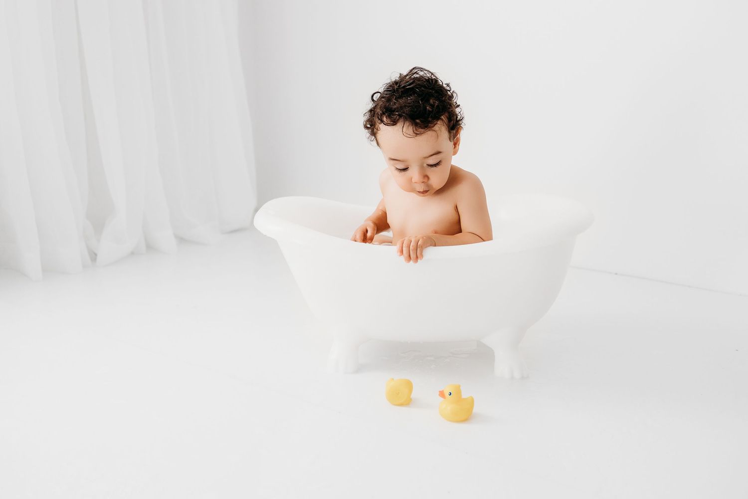 Baby boy with curly hair after a cake smash playing with yellow duckies. Portrait at his birthday photography session.