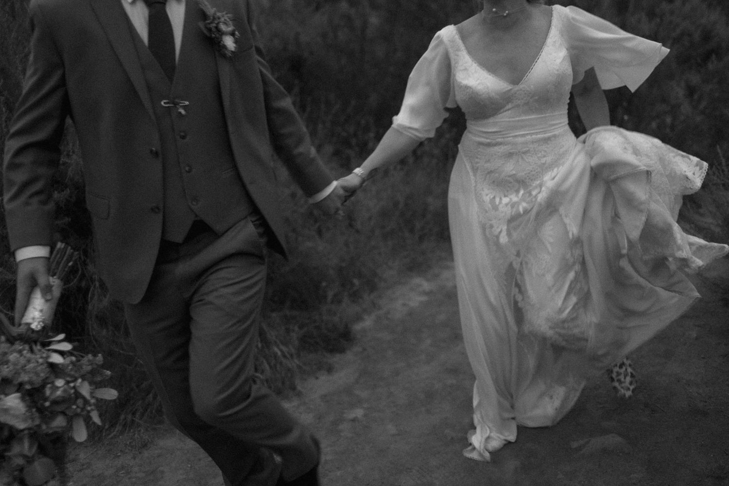 A black and white vintage-style photo of newlyweds running together on a path.
