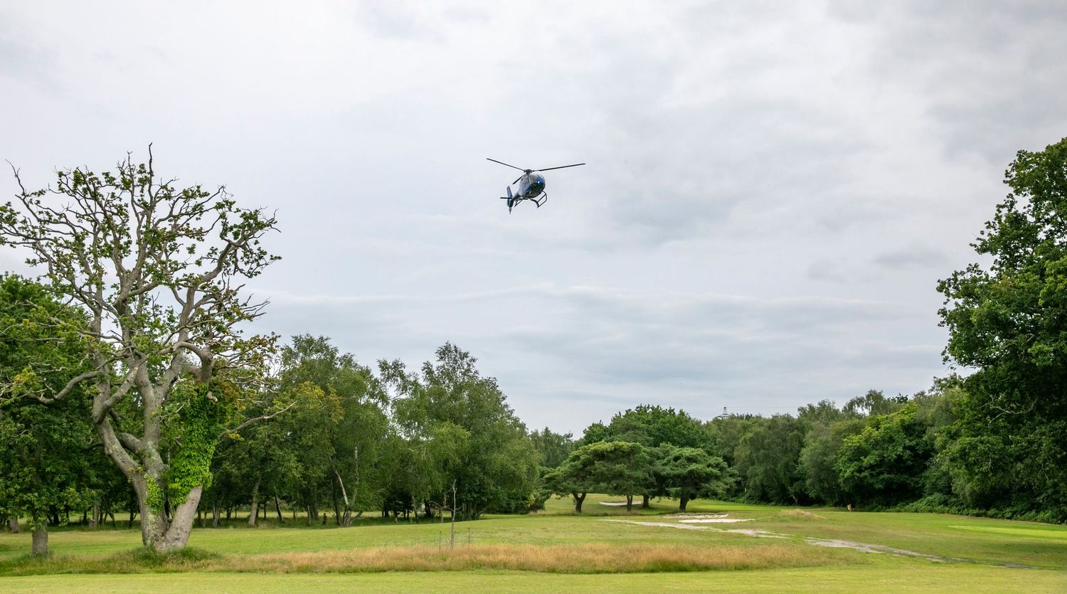 A helicopter flies over a scenic green golf course with scattered trees under cloudy skies.
