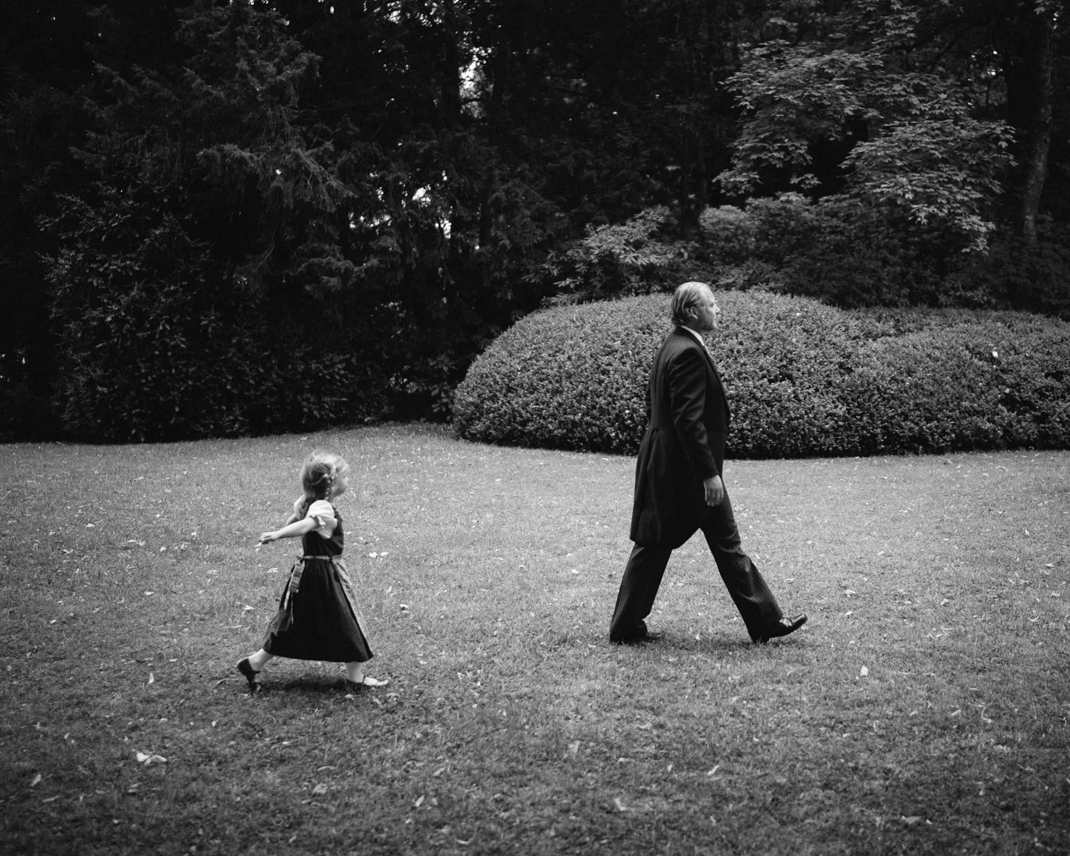 Black and white photograph of small child following adult across a garden lawn.