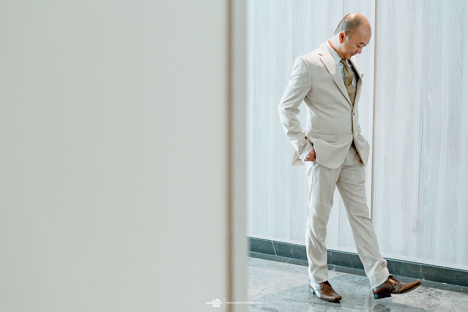 A person in a beige suit stands in an elegant pose against a minimalist white background.