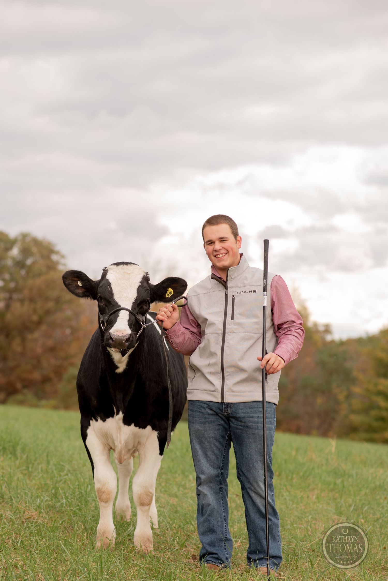 A Holstein dairy cow poses with handler holding staff in green pasture under cloudy sky.