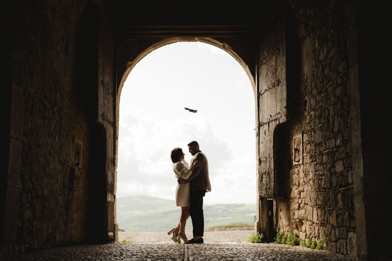 Silhouetted couple standing in a stone archway with a bird flying overhead.