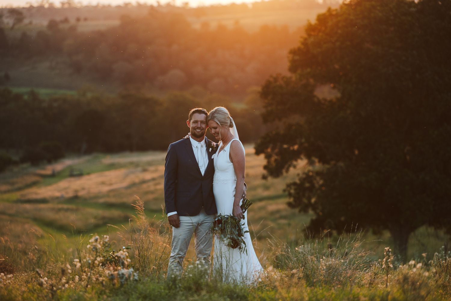 Bride and groom embrace at golden sunset overlooking lush green hills and wildflower meadows on their wedding day.