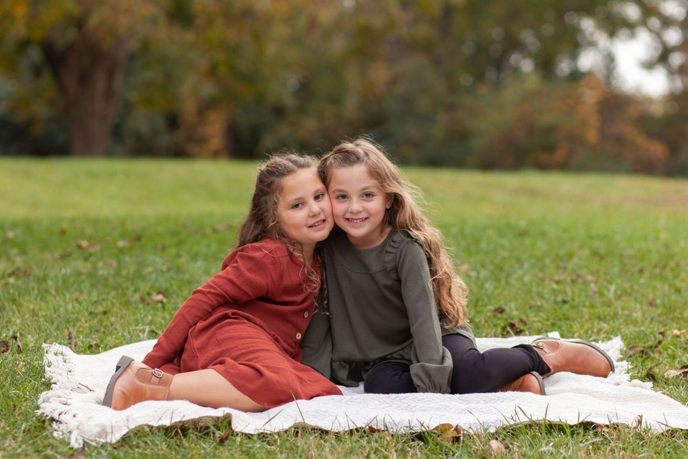 Two young children in autumn attire sit together on white blanket on grassy hill during fall photo session in Raleigh.