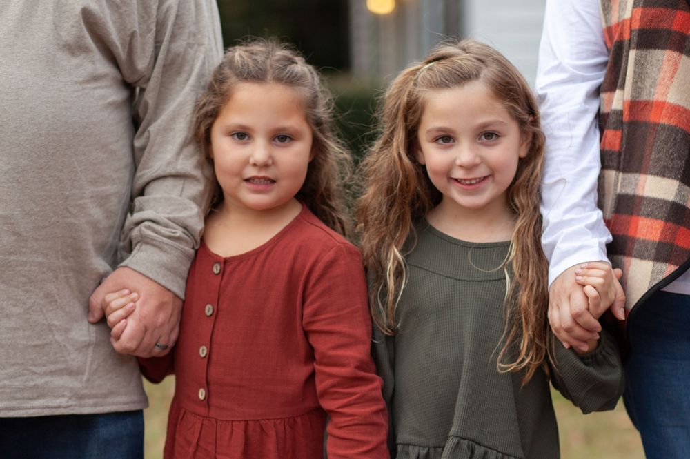 Two young children wearing autumn colored dresses hold hands with adults while standing together outdoors.