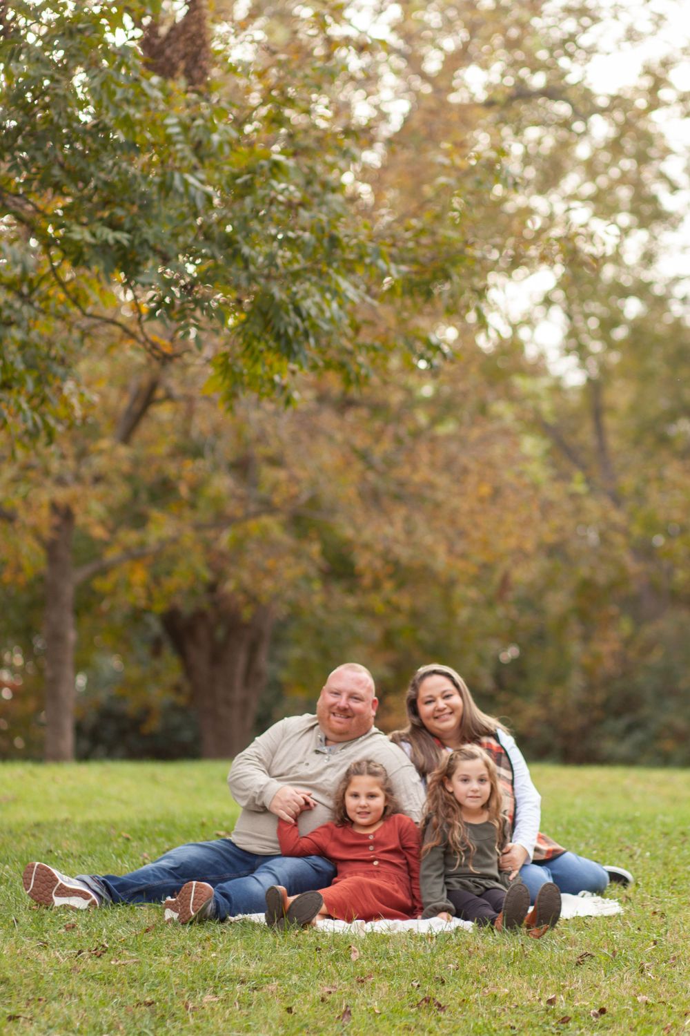Family sits together on grass in a park during autumn with fall foliage and trees in background at this Raleigh park.