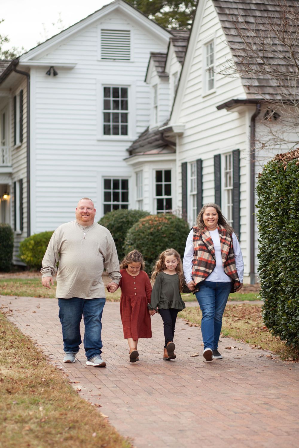 A family walking together on a brick pathway outside a classic white country house with black shutters at Historic Oak View Park in Raleigh