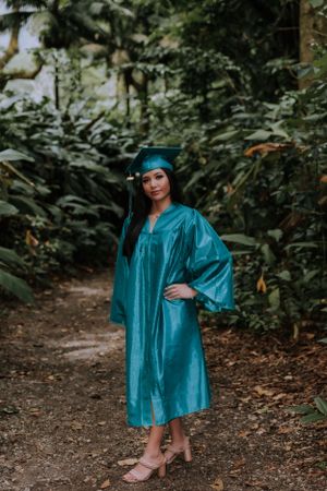 Full-length graduation portrait in teal cap and gown on garden path.