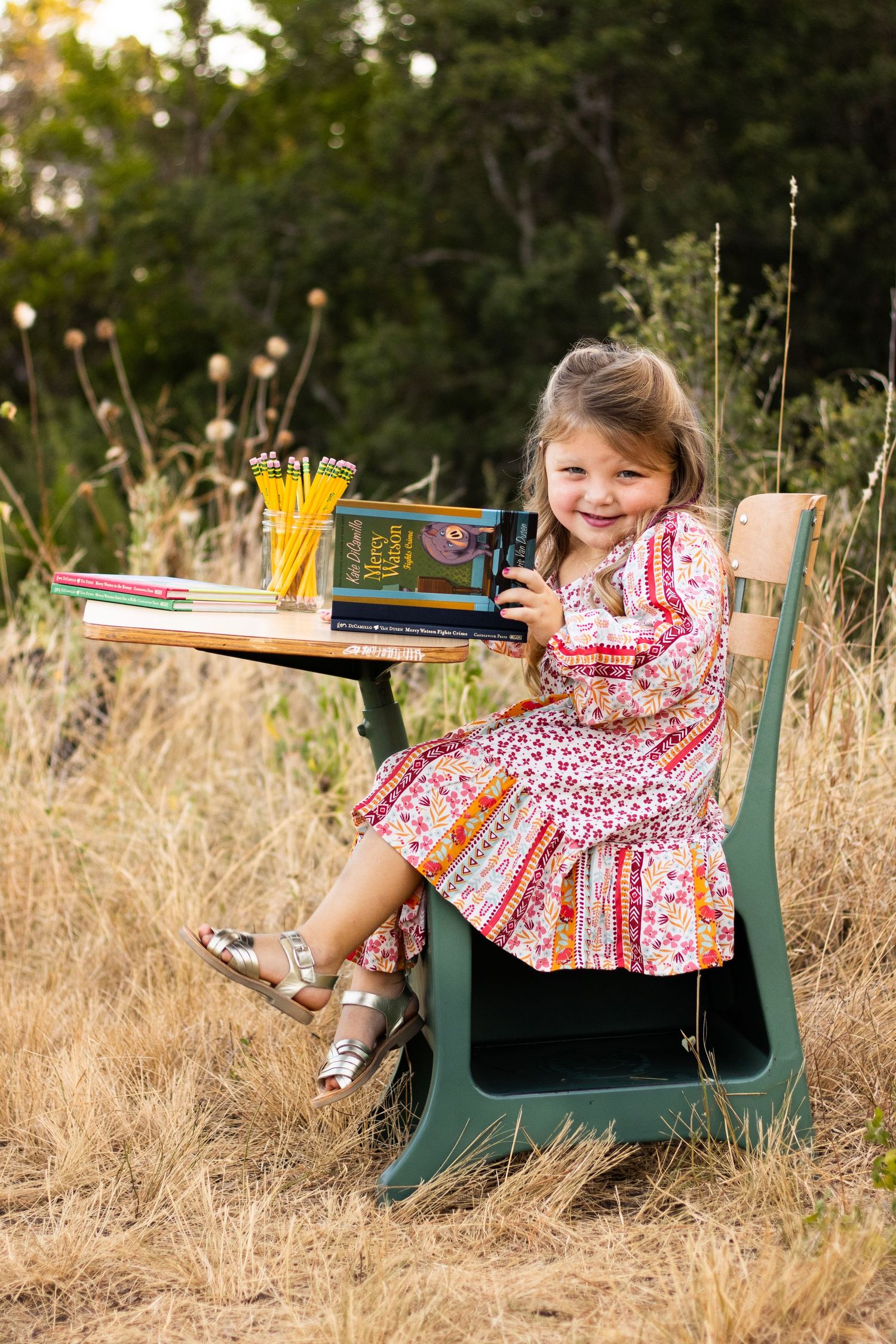 Series of shots showing young girl in patterned dress playing on green school desk outdoors in tall grass.
