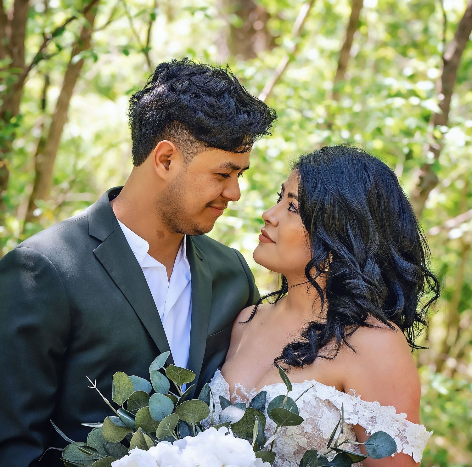 Couple in wedding attire sharing intimate moment with white bouquet in forest setting.