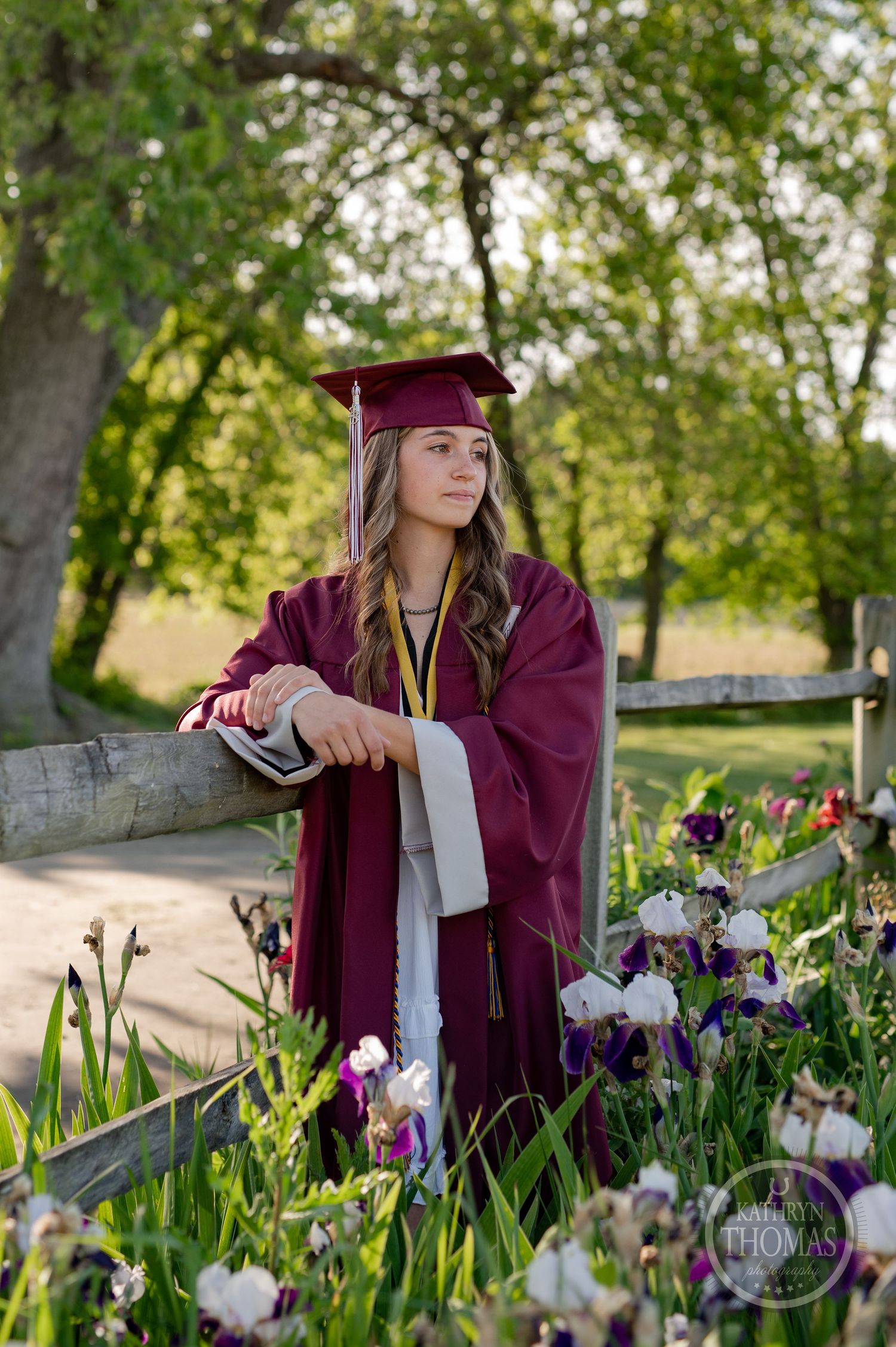 Graduate in maroon cap and gown poses by wooden fence surrounded by purple iris flowers.