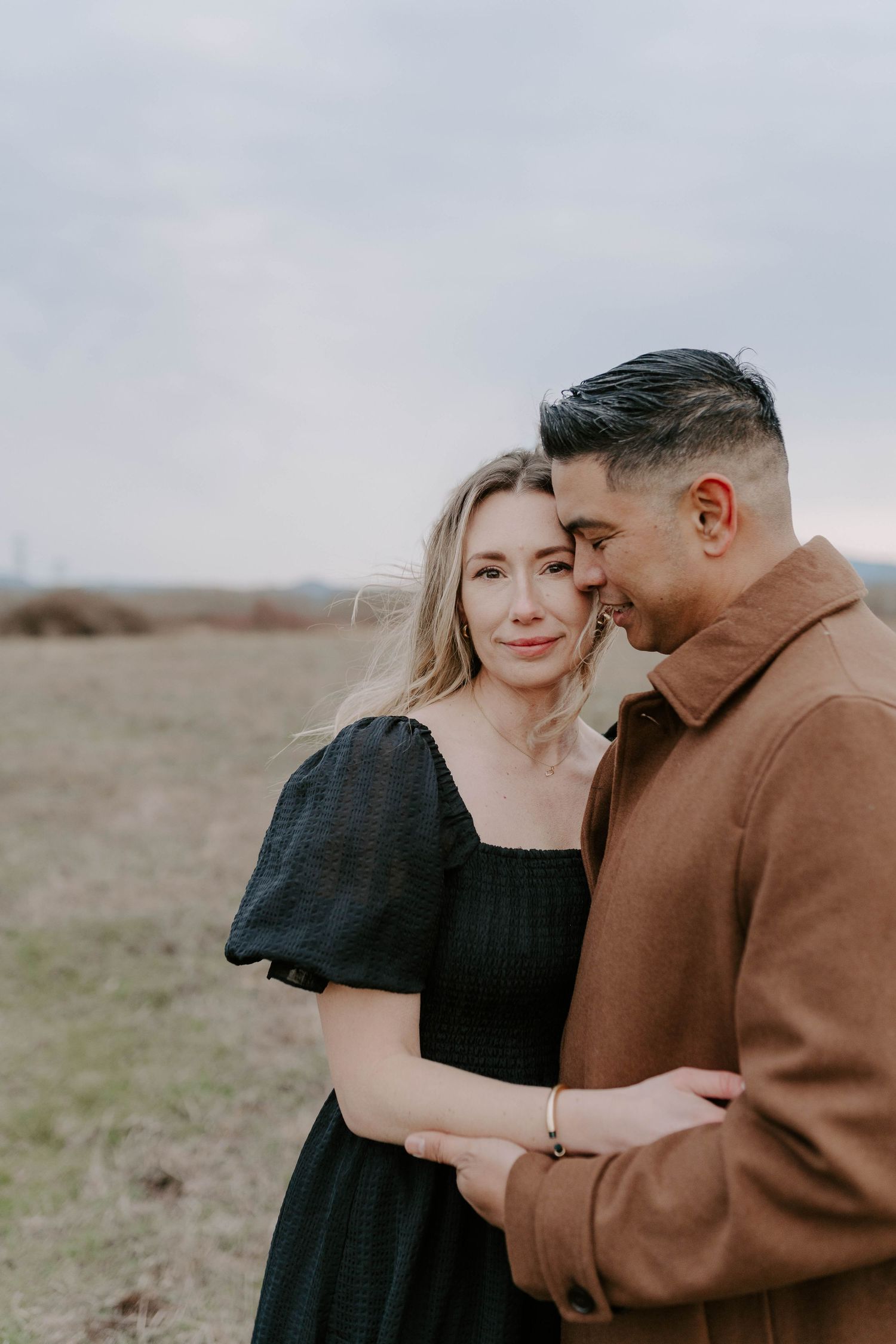 Romantic couple embracing in casual attire against overcast sky in open field.