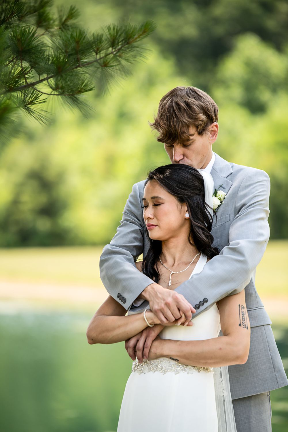 Tender embrace between newlyweds beside a tranquil lake with evergreen trees.