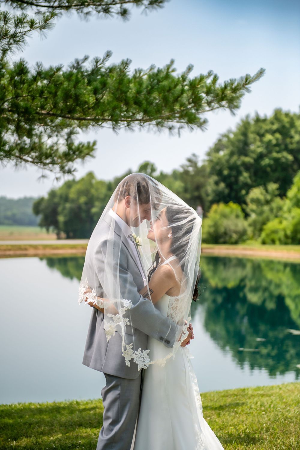 Intimate moment between newlyweds under veil by lakeside with pine trees overhead.