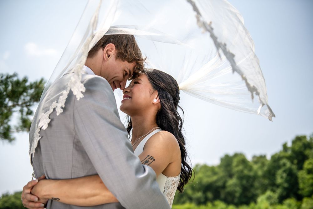 Romantic moment between newlyweds under flowing veil with natural backdrop.
