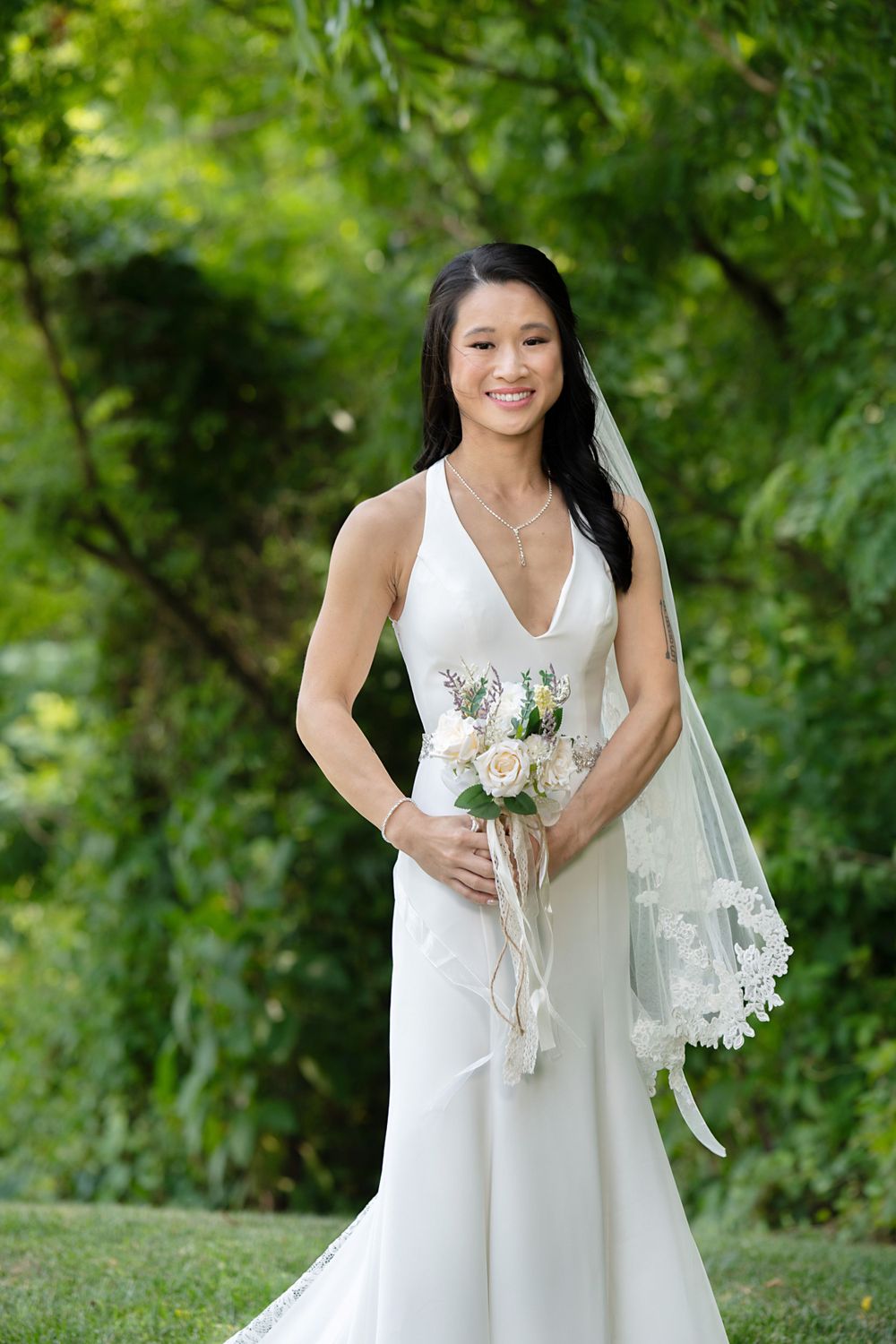 A bride in a white wedding dress holds a bouquet of white flowers against a lush green garden background.