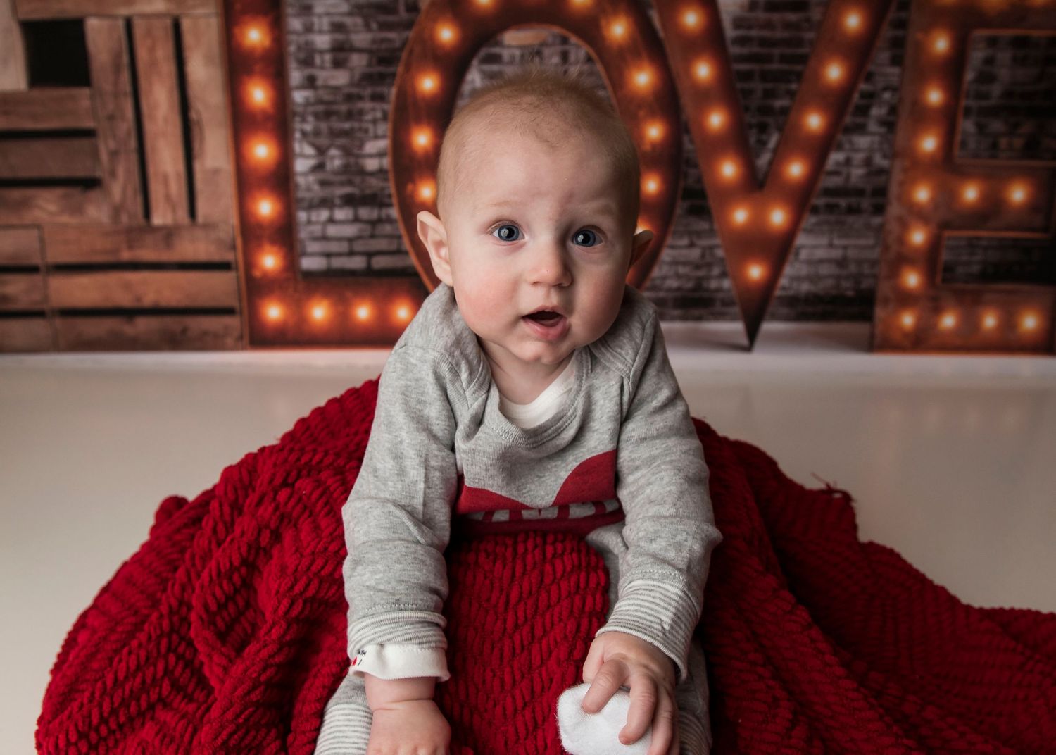 Baby in gray sweater sits on red blanket against brick wall with illuminated LOVE marquee letters.