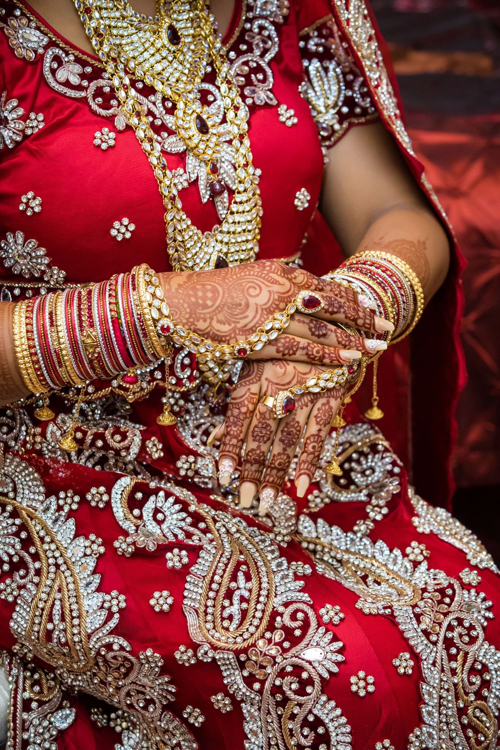 Detailed view of hands adorned with mehndi and bangles against red embroidered fabric.