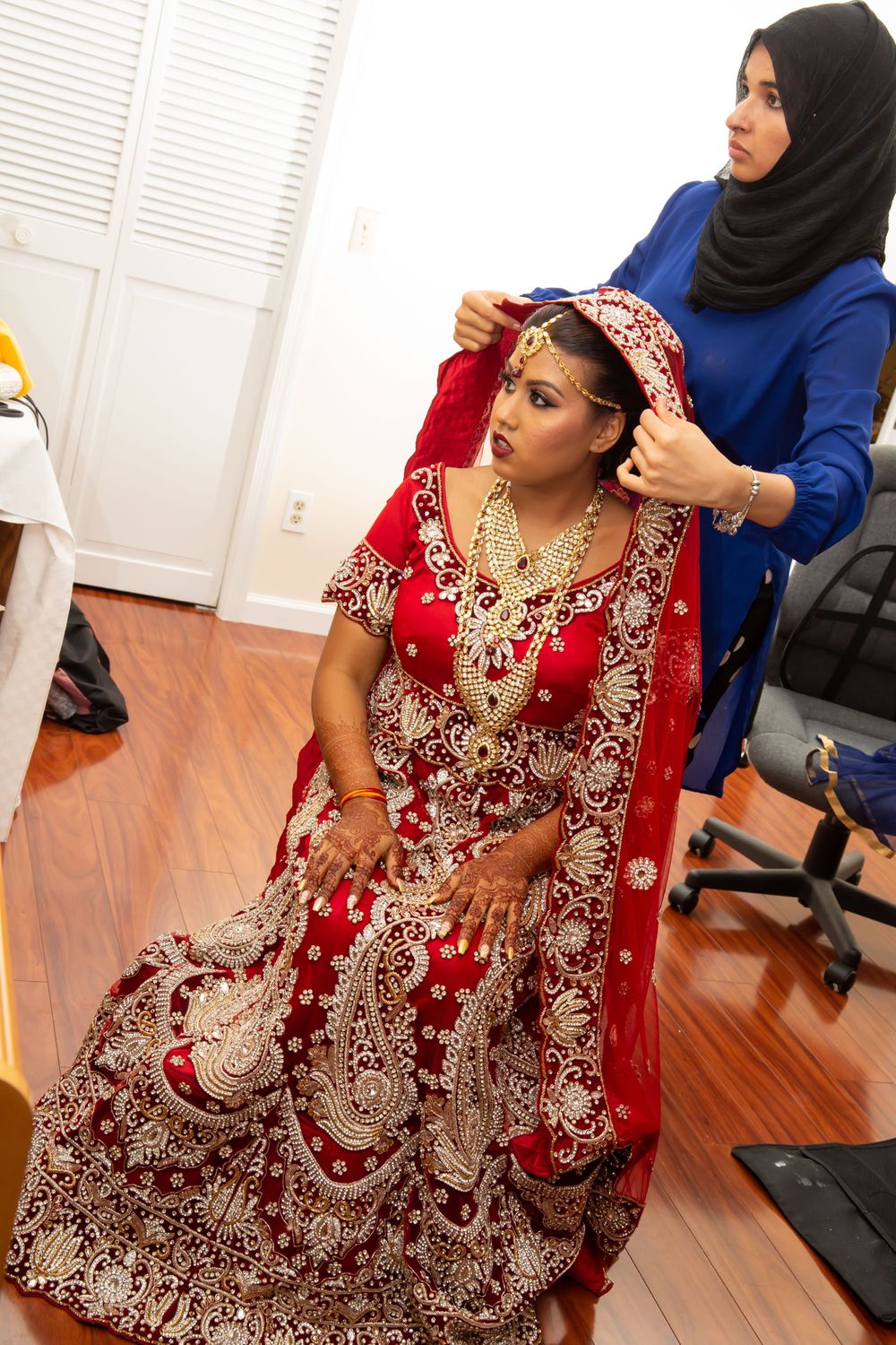 Traditional Indian bridal preparation scene with attendants helping adjust an embellished red lehenga.