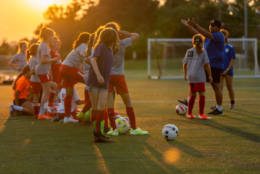 Soccer practice session at sunset with players gathered on field near soccer balls.