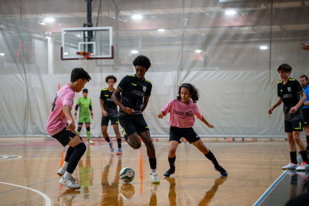 Youth indoor soccer players in pink and black jerseys compete for the ball on court.