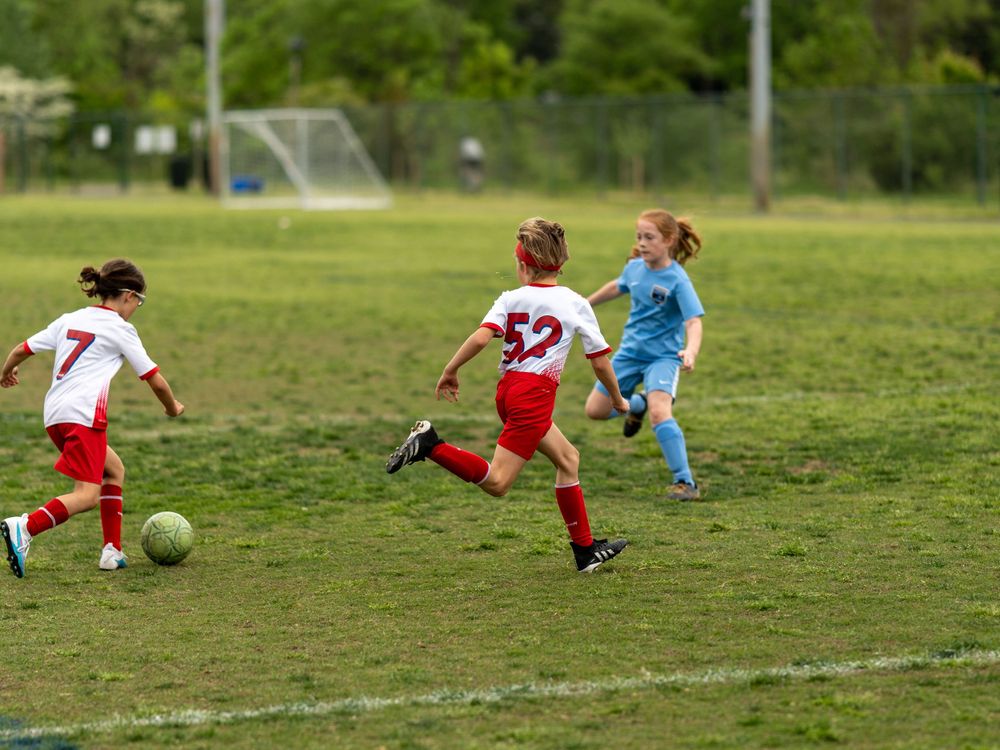 Youth soccer players compete for the ball on a grassy field with a goal visible in the background.