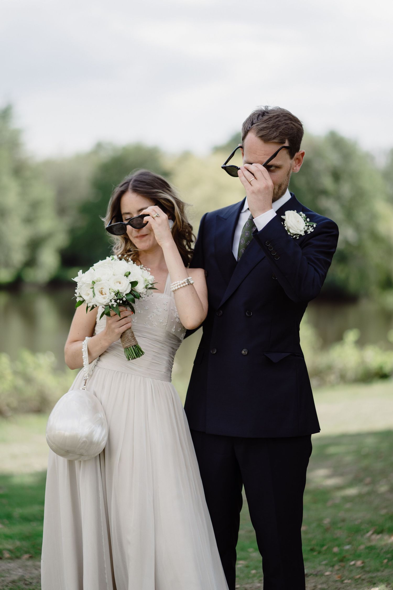 A wedding couple in formal attire laughing while covering their faces outdoors.
