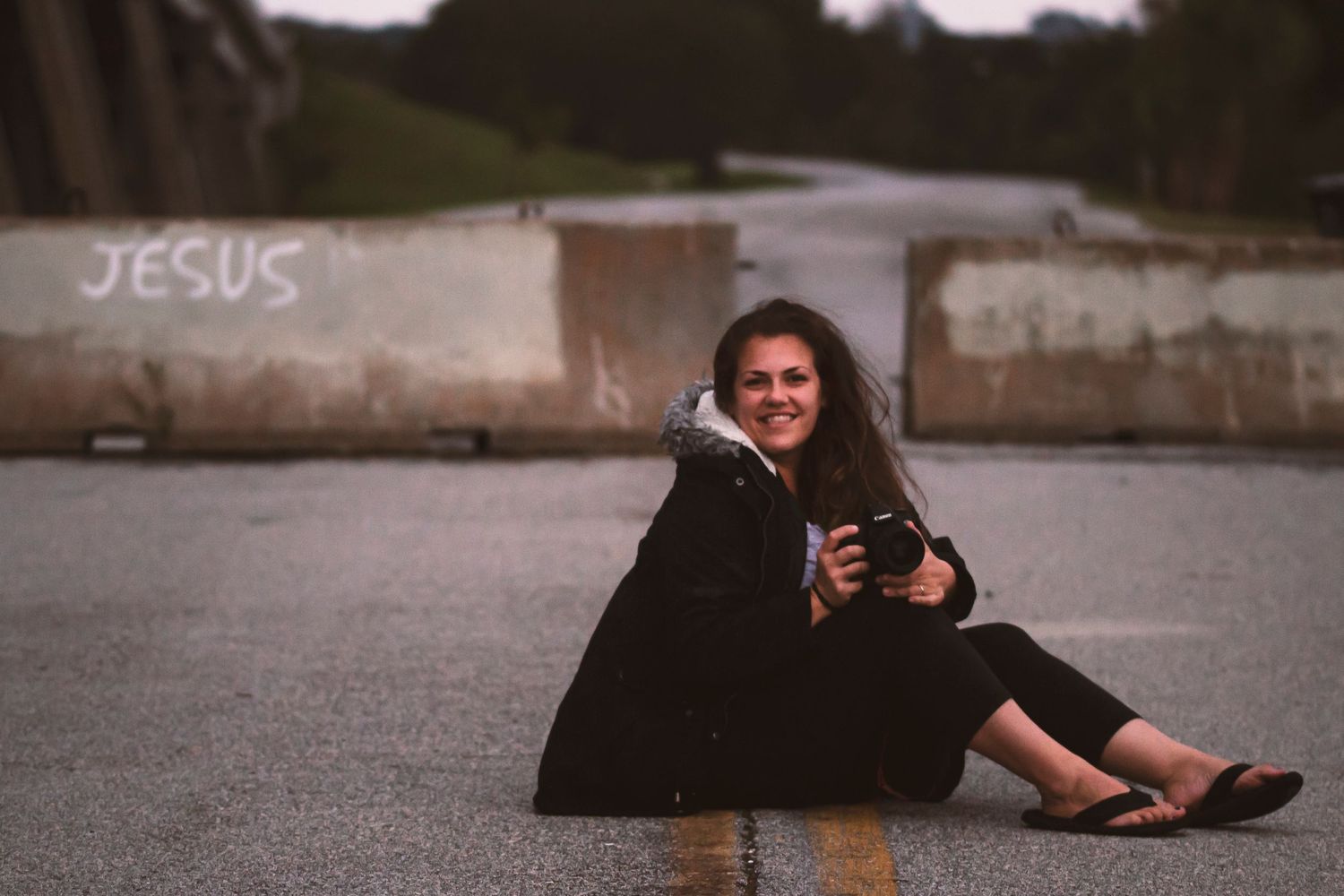 A photographer sits on pavement with camera near graffiti wall at dusk.