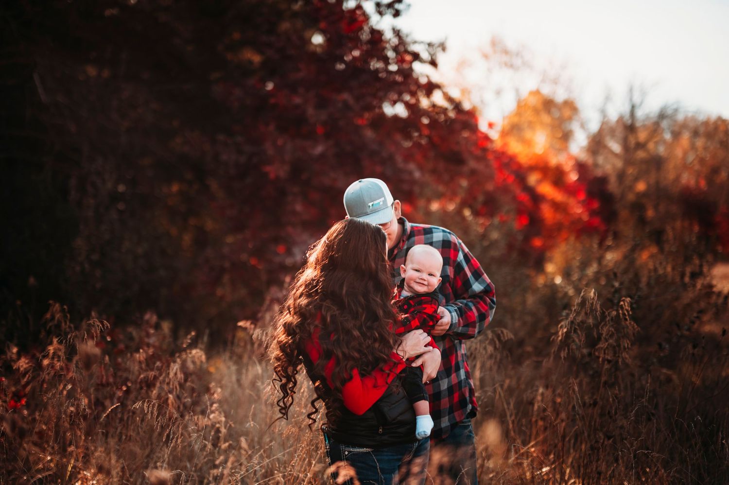 Parents sharing a kiss while holding their baby in a vibrant autumn field.