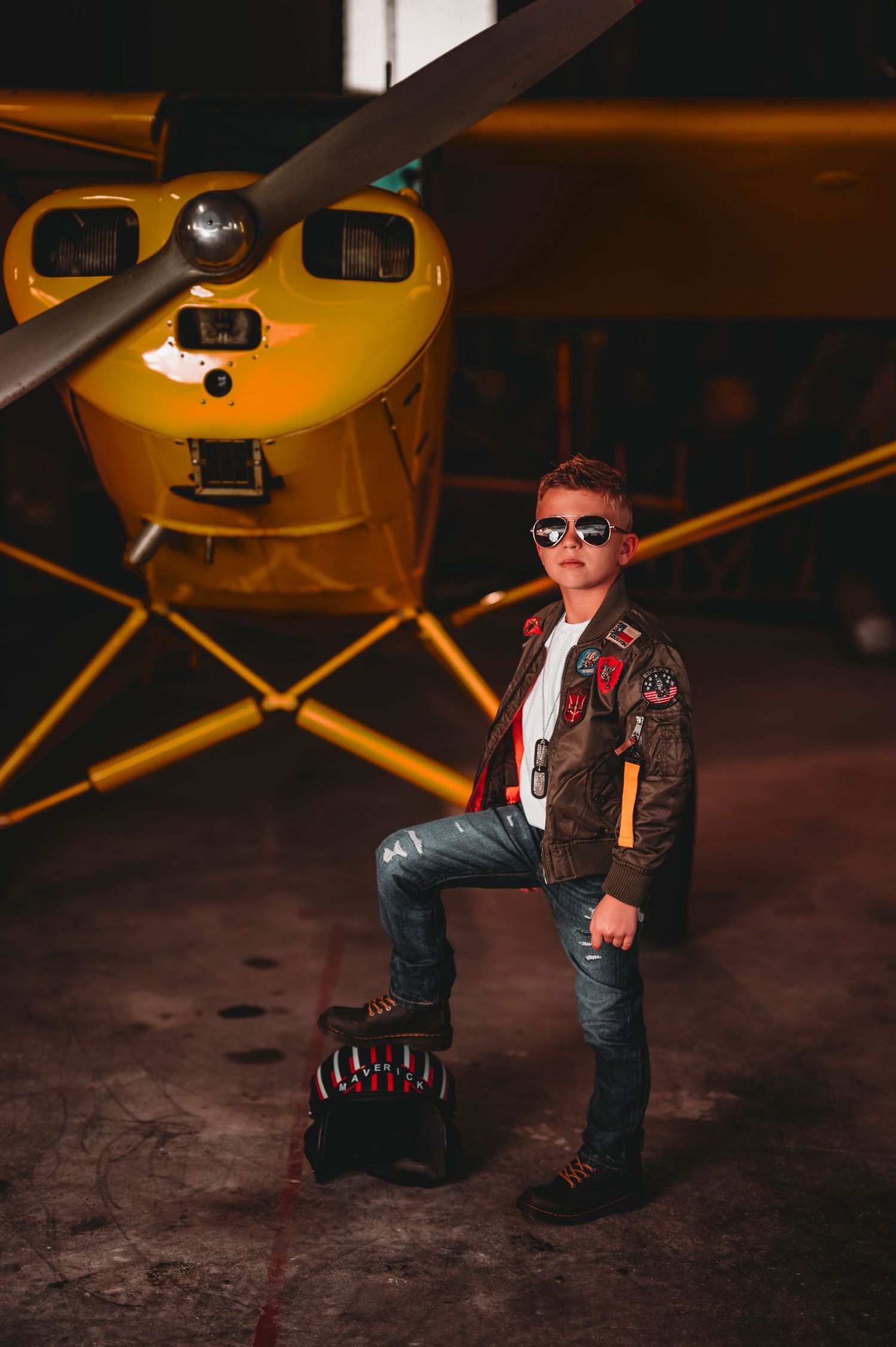 Cool young boy in aviator jacket and sunglasses posing beside a yellow plane.
