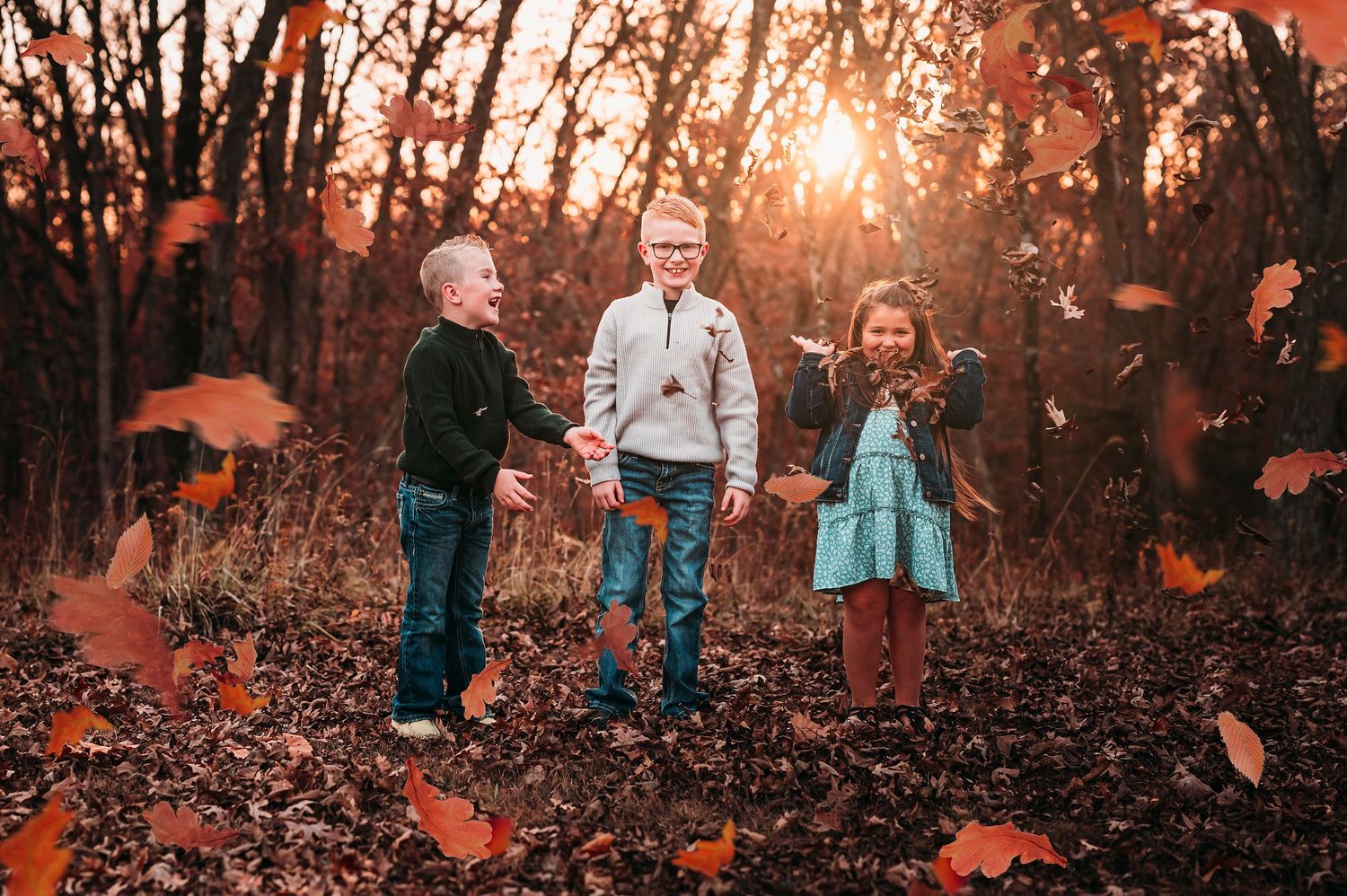 Three children playing in autumn leaves in a forest with golden sunlight filtering through red and orange trees.