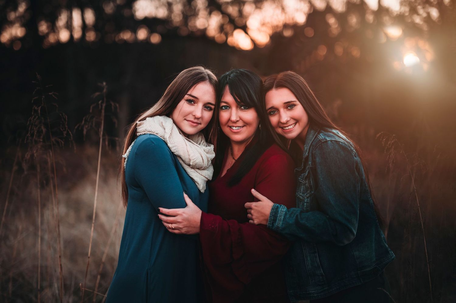 Three women smiling and hugging warmly in a sunlit golden field.