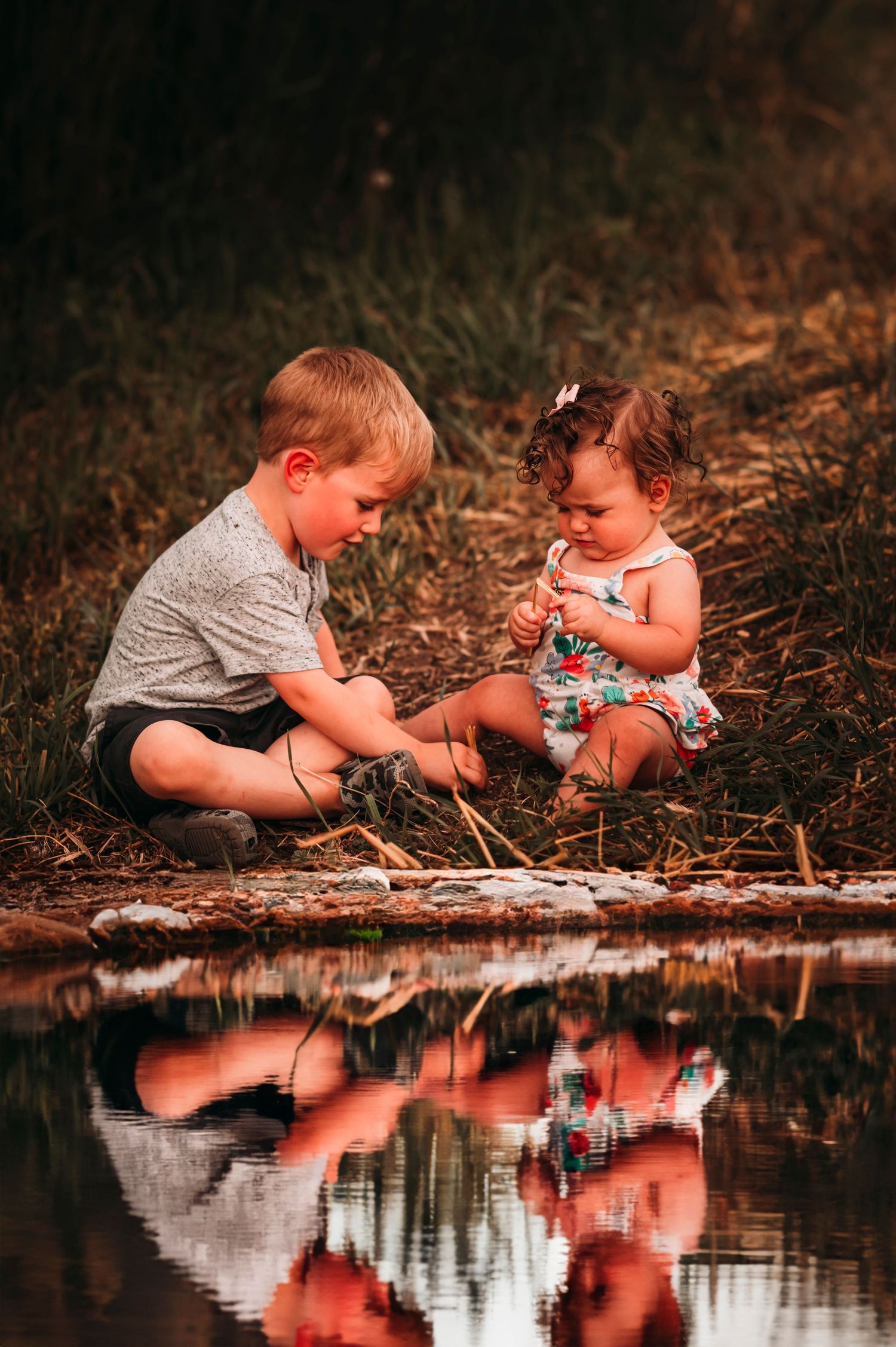 Young boy and baby girl sitting by a reflective pond playing with rocks in a serene outdoor setting.