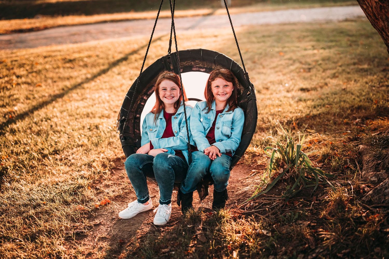 Two young girls in matching outfits smiling while sitting together on a tire swing at sunset.