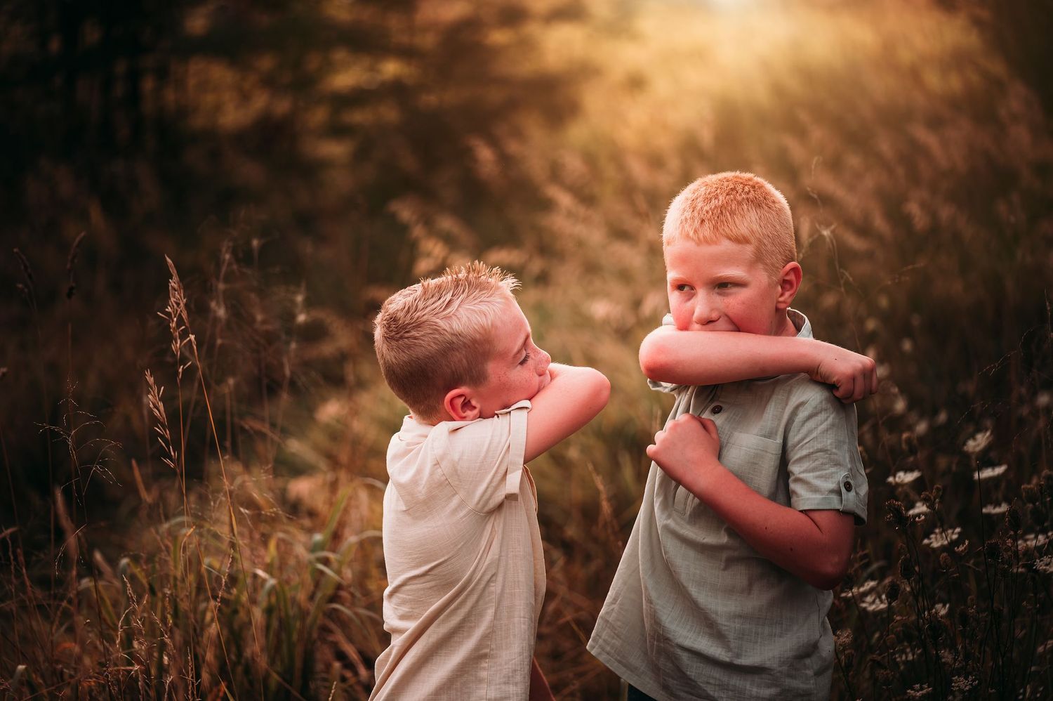 Two young blonde boys hugging each other playfully in a sunlit grassy meadow.