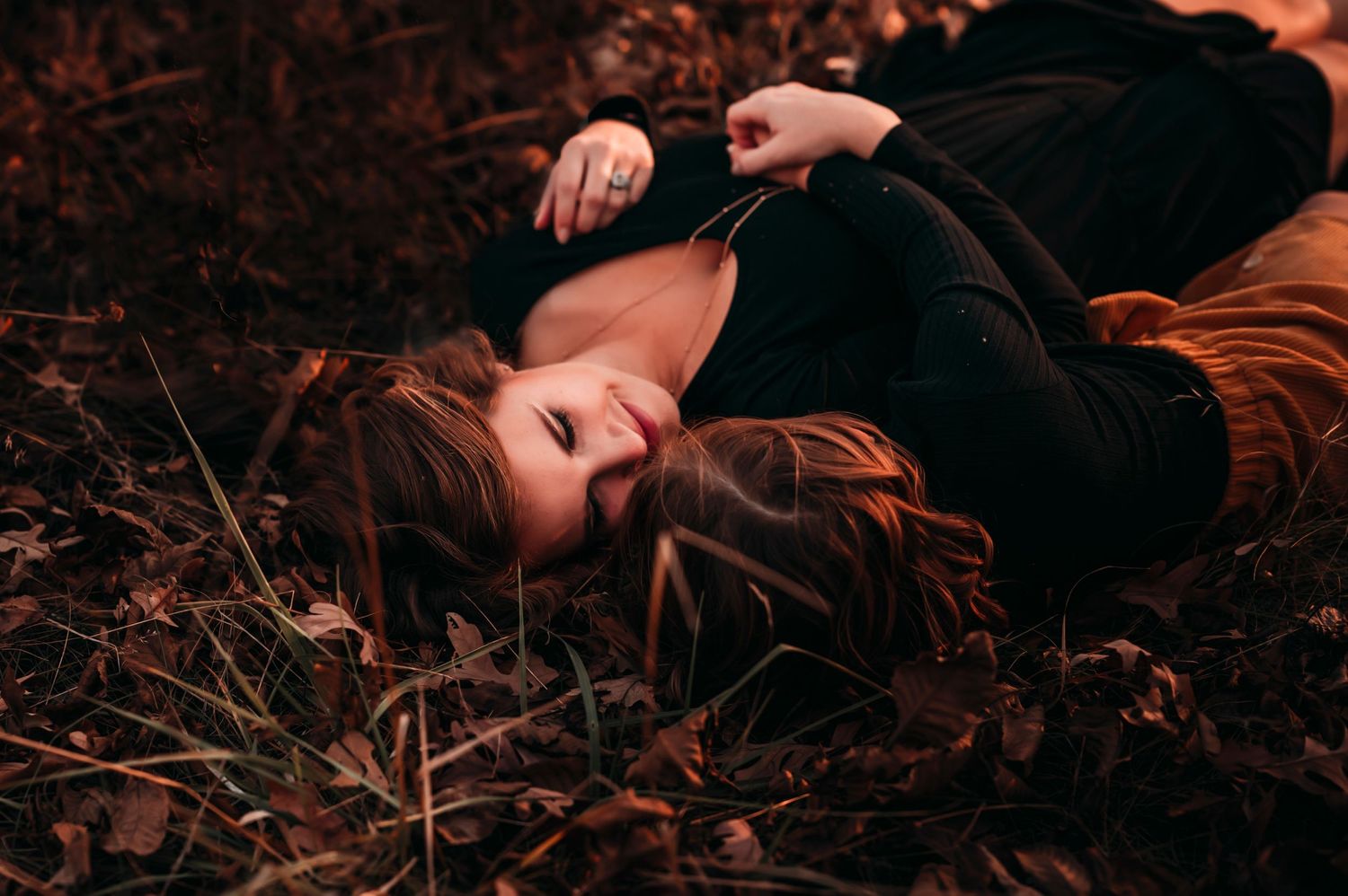 Young woman with long hair lying peacefully among fallen autumn leaves and grass in a moody outdoor setting.