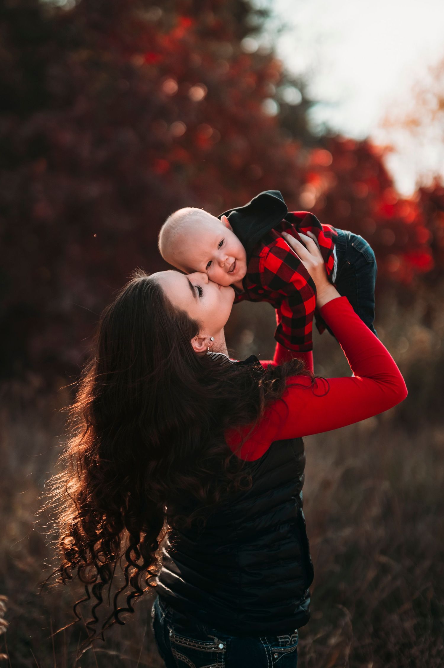 Mother joyfully lifting and kissing her smiling baby in a red autumn setting.