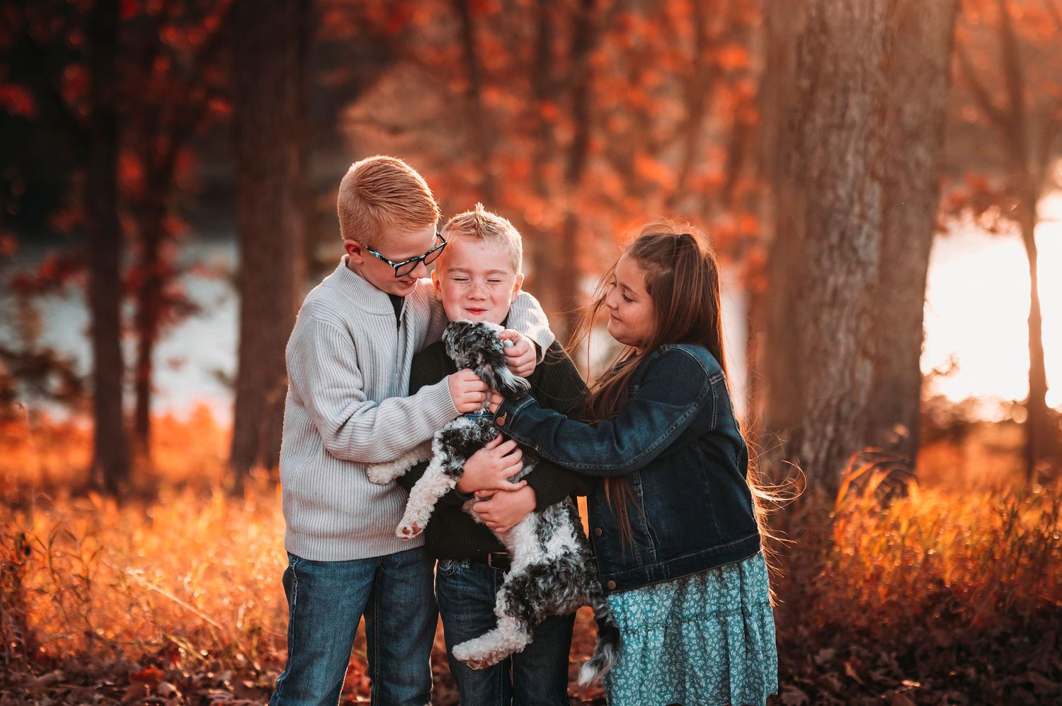Two children and a teenage girl cuddling a small dog in a stunning autumn forest with warm golden light.