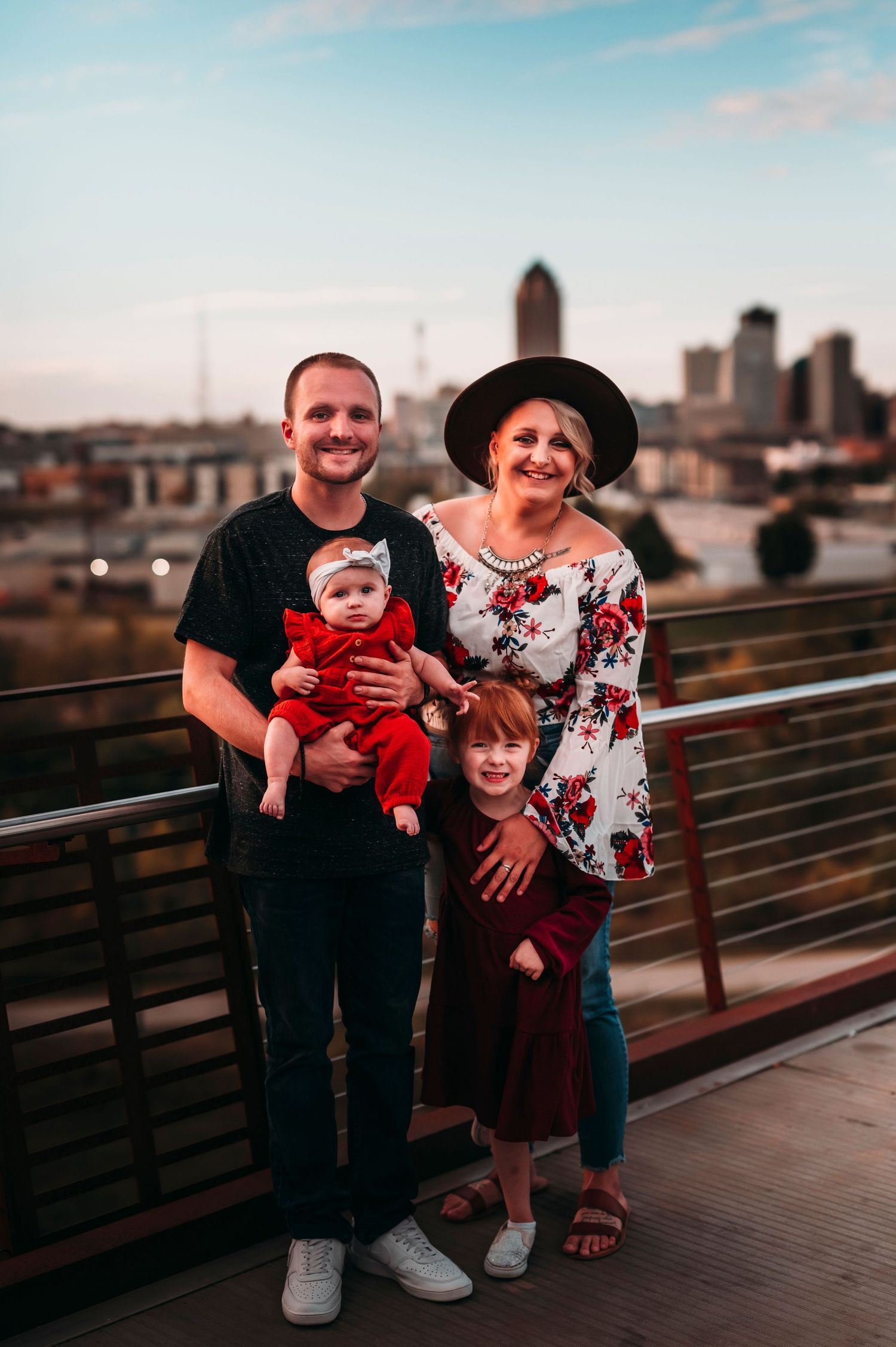 Happy family of four posing on a rooftop bridge with a city skyline at dusk.