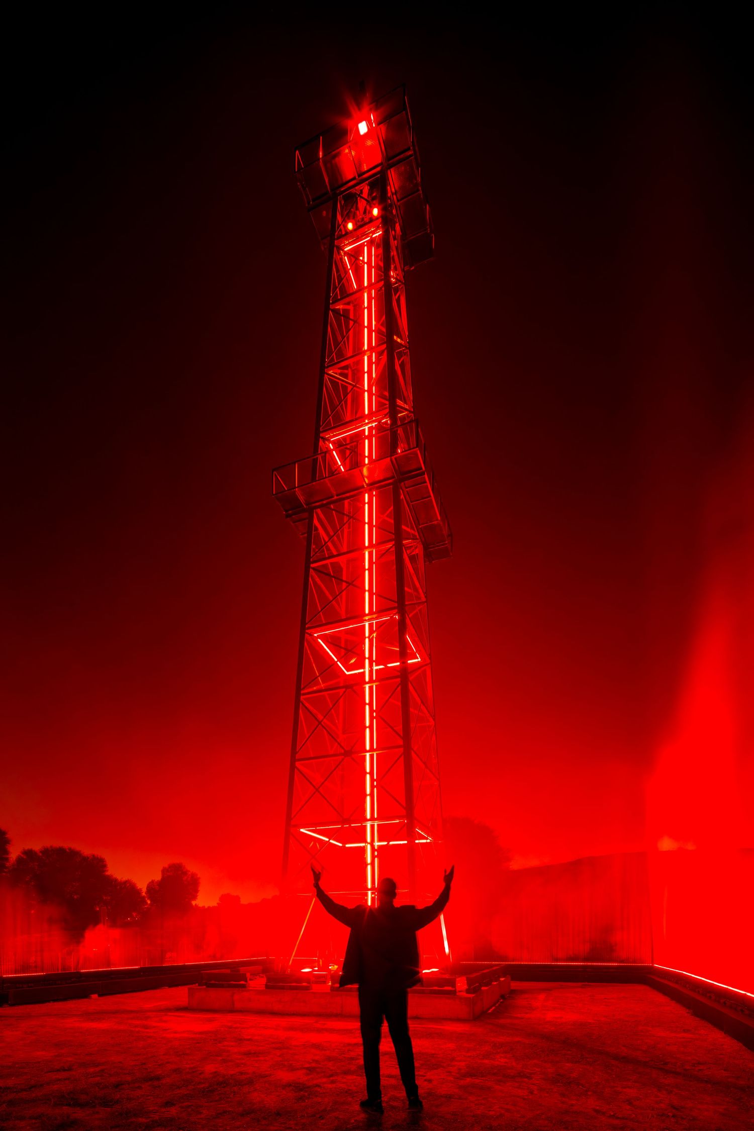 Tall transmission tower glows red against dark sky with silhouetted figure in foreground.