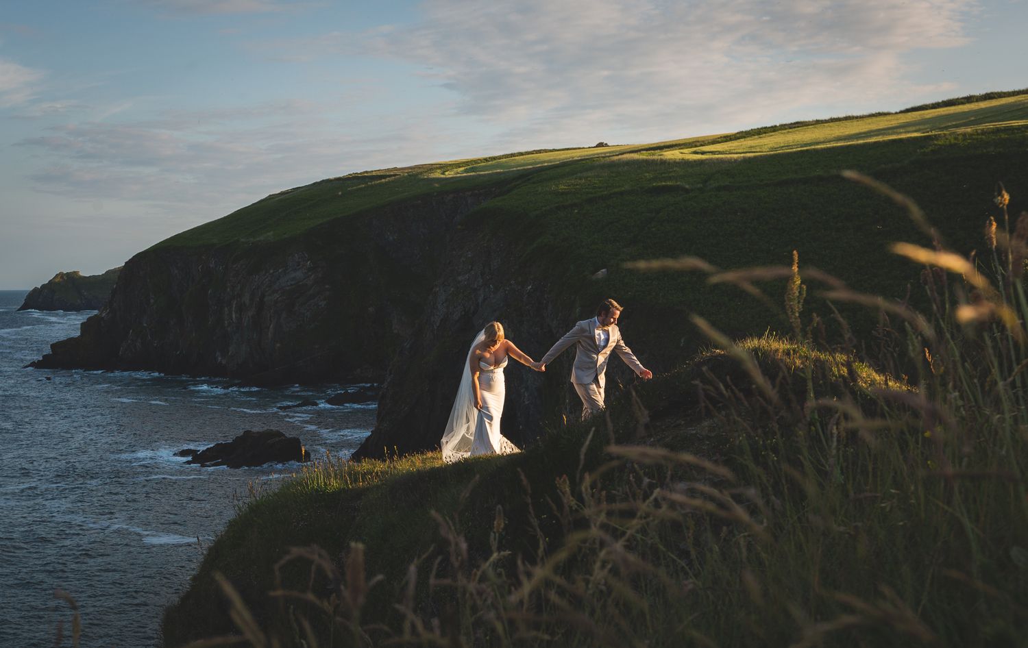 Two figures walk along dramatic coastal cliffs at sunset with ocean waves crashing below.