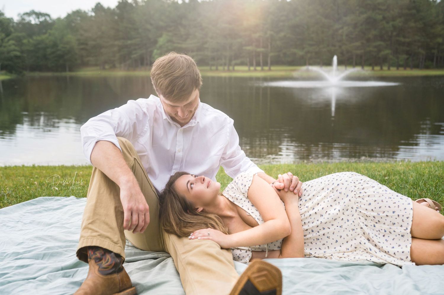 Couple lounging on picnic blanket by lake with fountain in background.
