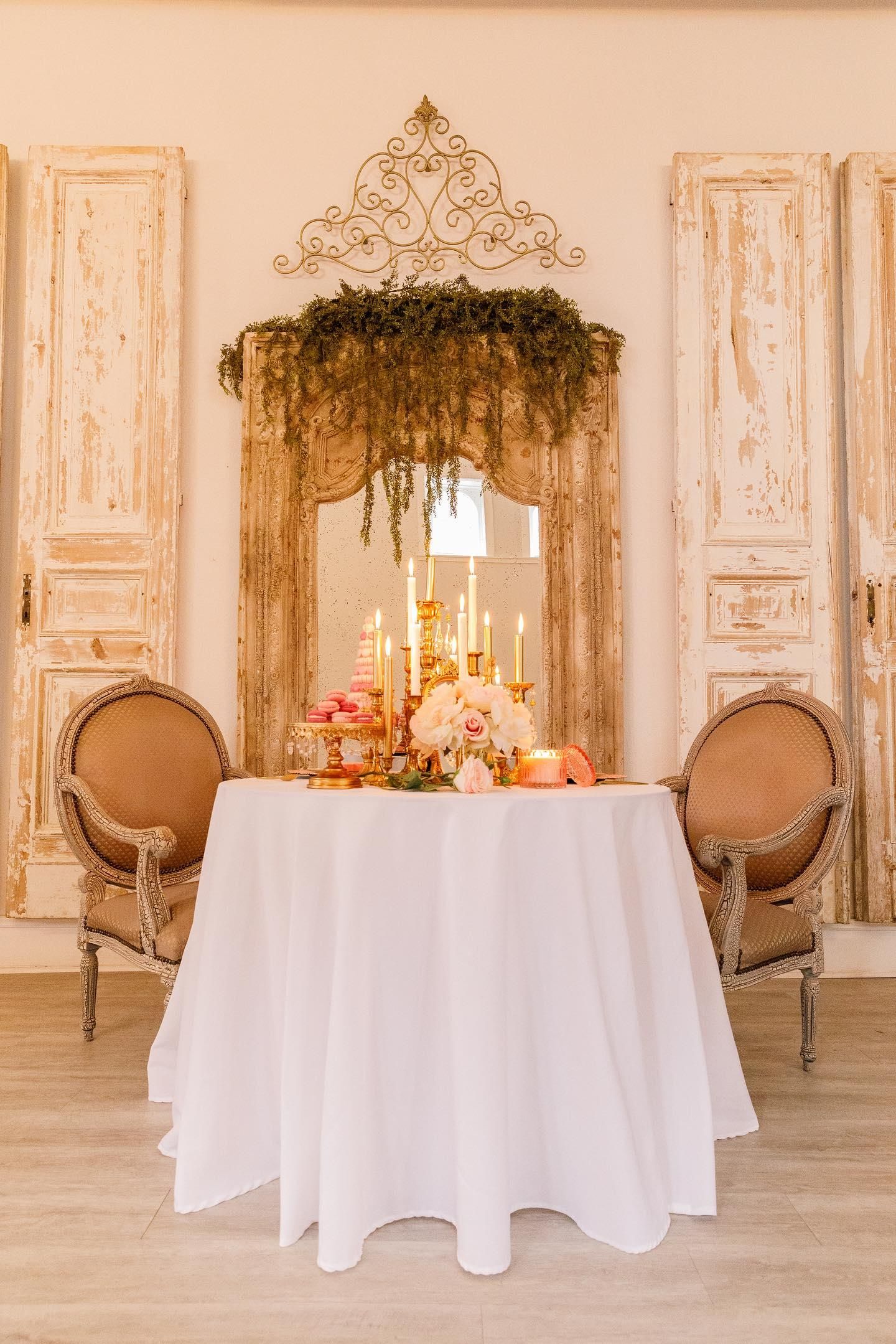 Intimate dining setup with white tablecloth, vintage chairs, and ornate doorframe decorated with cascading greenery.