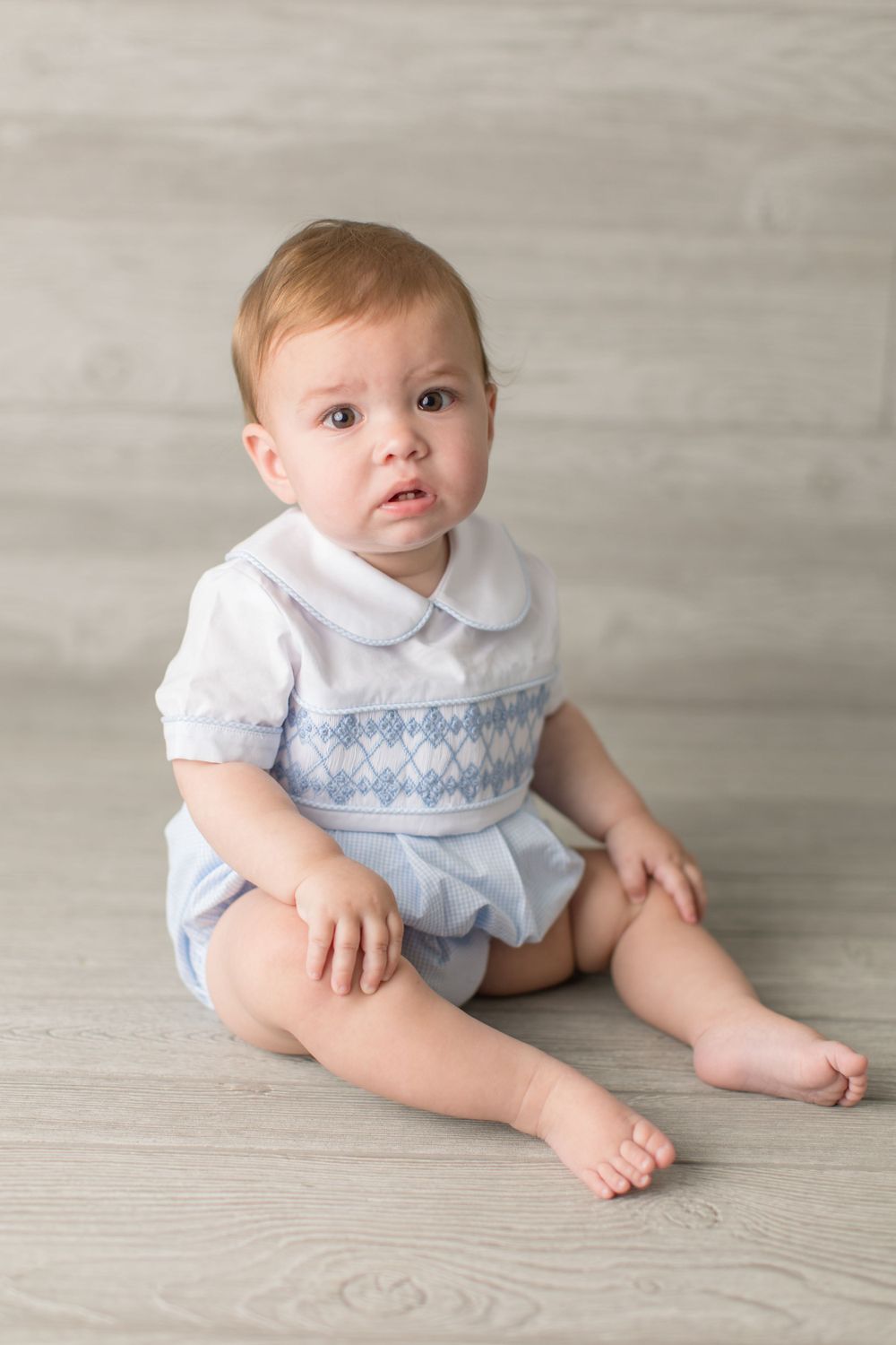 Small child in blue and white smocked outfit sitting on light wooden floor.