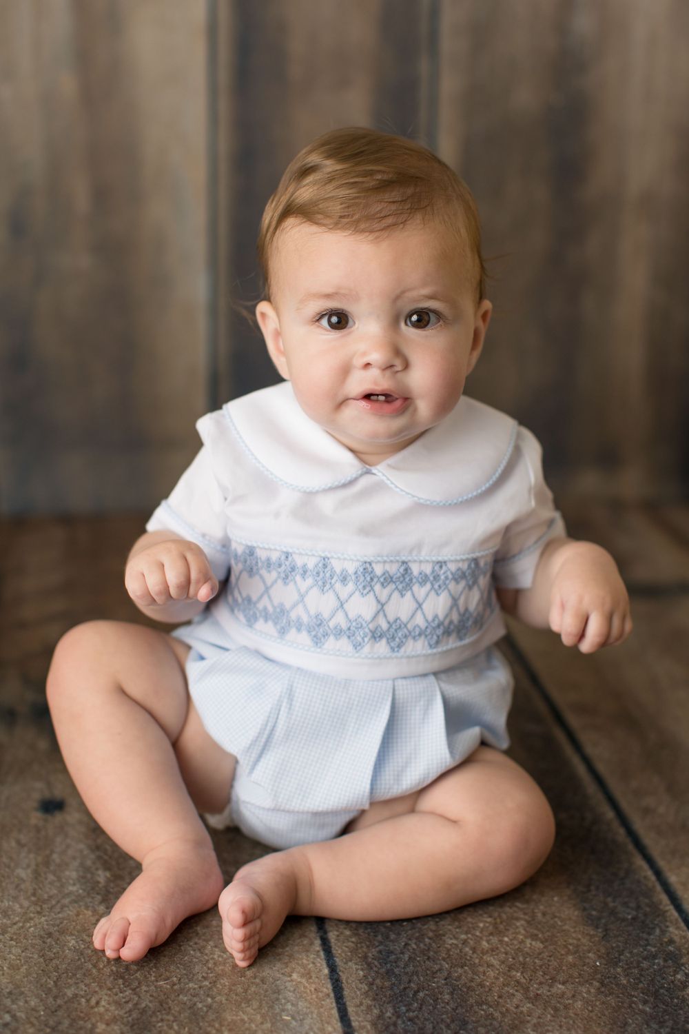 Baby wearing smocked white and blue outfit sitting on wooden floor.