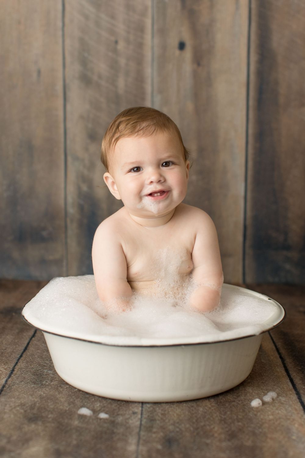 Baby sitting in vintage white basin with bubbles against wooden backdrop.